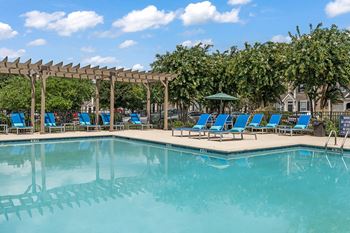 A swimming pool with blue chaise lounge chairs and a pergola with trees in the